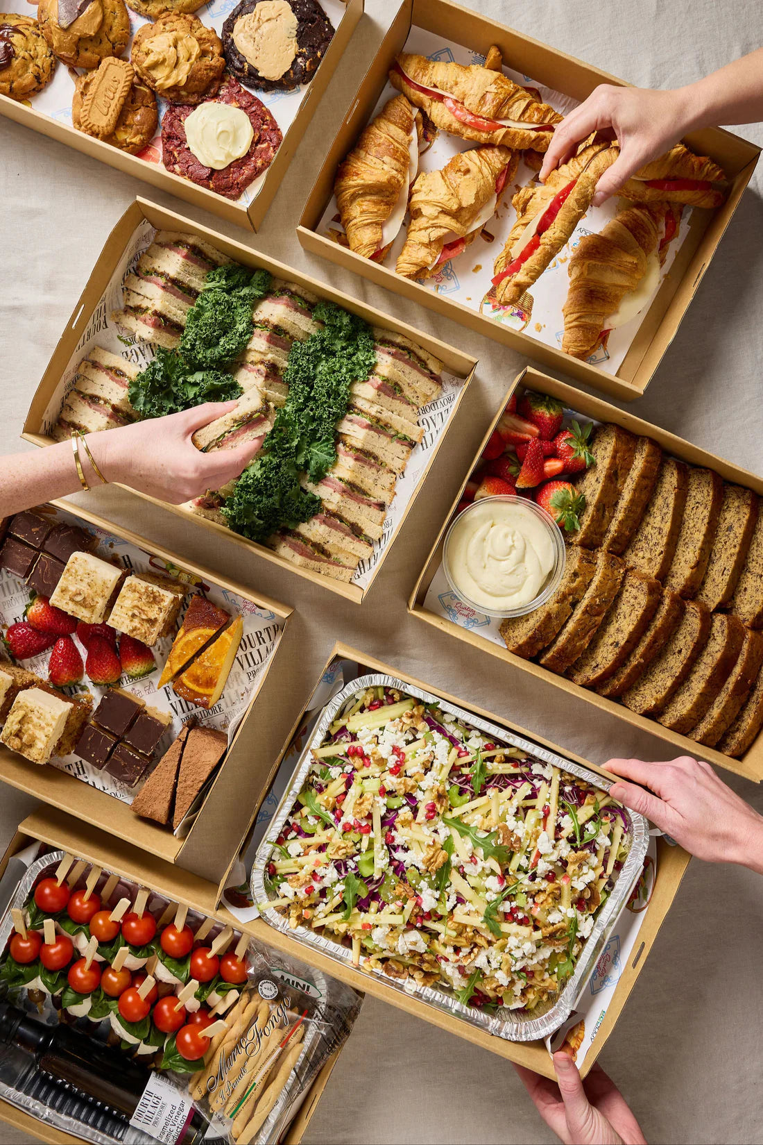 Assorted catering including sandwiches, salads, and desserts in a wooden box on a neutral background.