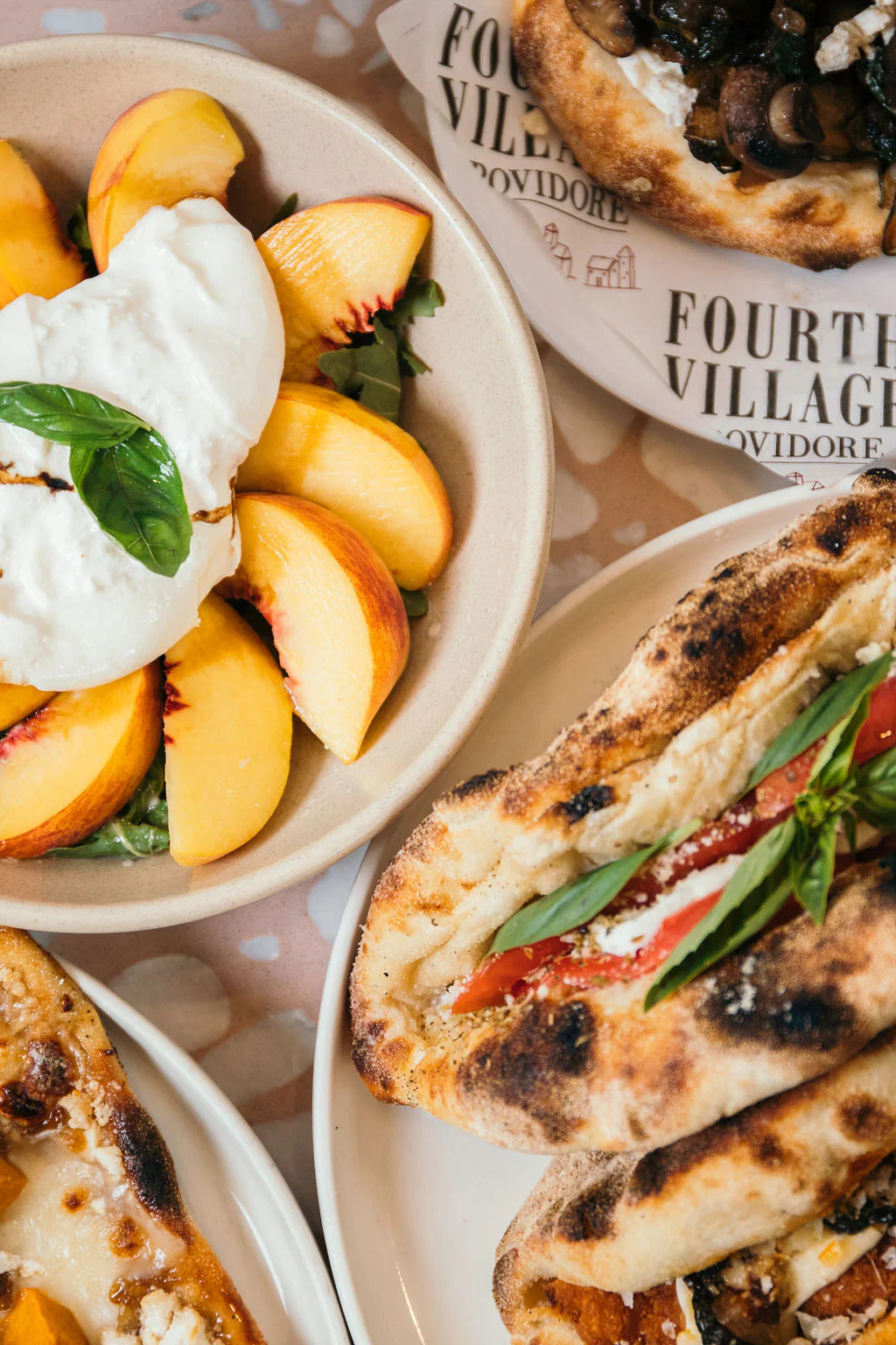 Plates of food including a salad with peaches and burrata, and grilled sandwiches with vegetables on a table.